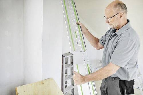 Une personne portant un t-shirt gris utilisant un outil Festool près d'un mur, en ajustant un rail-guide. Une planche de bois est située à gauche.