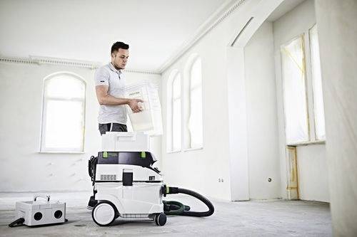 Un homme en chemise blanche charge un aspirateur blanc dans une pièce lumineuse, vide, avec de grandes fenêtres, suggérant un nettoyage ou un déménagement.