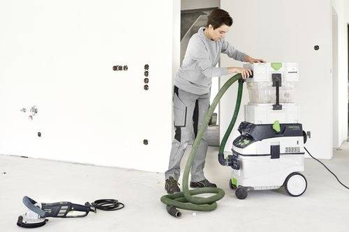 Une personne en tenue de travail ajuste un tuyau sur un grand aspirateur industriel dans une pièce non meublée. Un autre outil électrique est posé au sol.