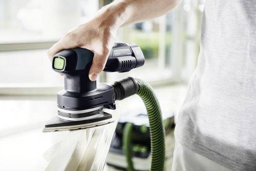 Une personne utilise une ponceuse électrique portable avec un tuyau d'aspirateur vert connecté, en lissant les bords du bois à l'intérieur, ce qui suggère des travaux de rénovation domestique.