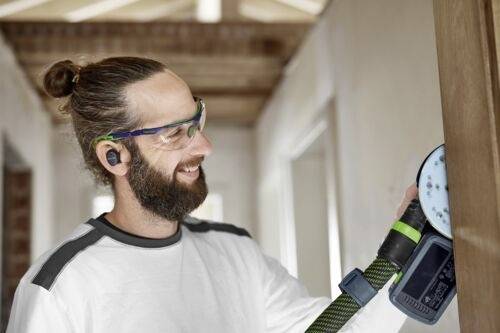 Un homme avec une barbe et des lunettes de sécurité utilise une ponceuse sur une surface en bois à l'intérieur, ce qui indique une activité de rénovation domiciliaire.