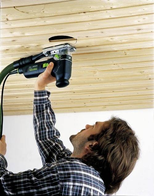 Une personne utilise un outil électrique pour percer un trou dans un plafond en bois, l'outil étant relié à un tuyau pour la collecte des poussières.