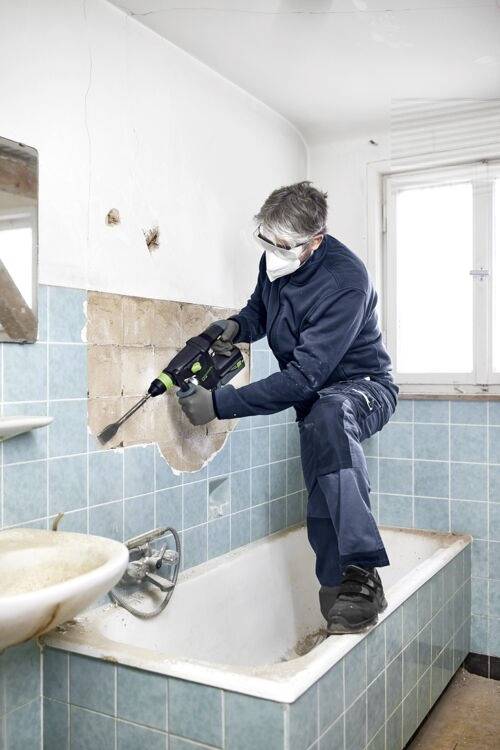 Une personne portant un équipement de protection rénove une salle de bain, utilisant un outil électrique pour enlever des carreaux d'un mur au-dessus d'une baignoire.
