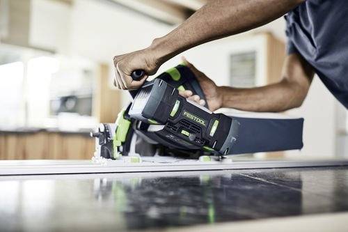 Une personne utilise une scie circulaire Festool verte et noire sur une surface en bois à l'intérieur, en se concentrant sur une coupe précise.