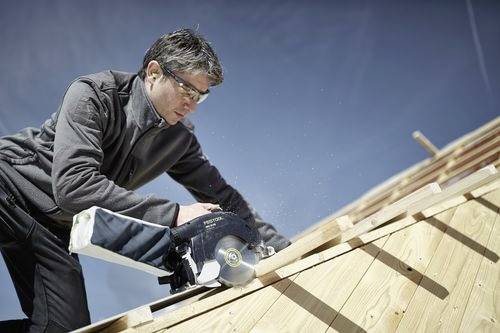 Une personne utilisant une scie circulaire pour couper des planches de bois sur un toit en pente, portant des lunettes de sécurité et des vêtements d'extérieur sous un ciel dégagé.