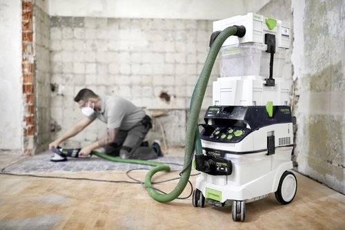 Une personne portant un masque ponce un sol en bois dans une pièce partiellement rénovée. Un extracteur de poussière est bien visible au premier plan.