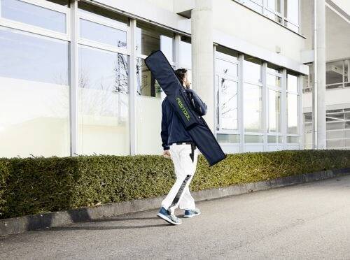 Une personne marche à l'extérieur d'un bâtiment moderne, portant un long sac noir sur l'épaule, suggérant un mouvement et un but.