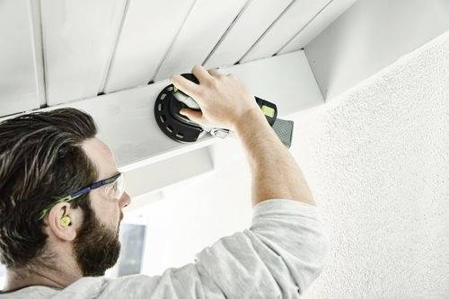 Une personne portant des lunettes de protection et des bouchons d'oreilles, ponçant une poutre de plafond avec une ponceuse électrique, concentrée sur une tâche de rénovation domiciliaire.