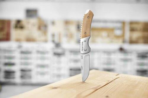 Un couteau pliant avec un manche en bois et une lame en acier inoxydable est dressé verticalement sur une surface en bois, avec un arrière-plan d'atelier flou.