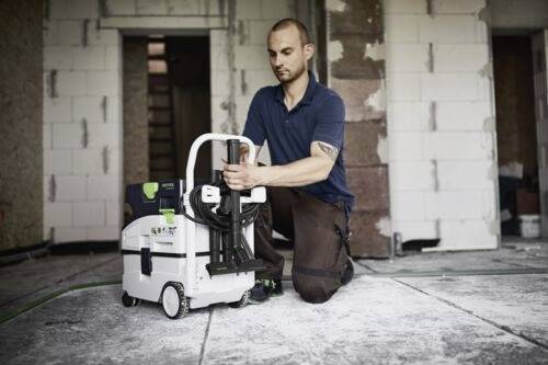 Une personne s'agenouille sur un sol de chantier, en ajustant une petite machine de nettoyage dotée d'une poignée et de roues. L'arrière-plan montre des murs non finis.