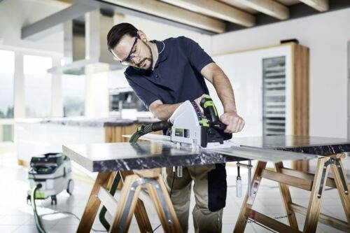 Une personne portant des lunettes de sécurité utilise une scie circulaire pour couper une grande planche sur deux tréteaux dans un atelier lumineux et moderne.