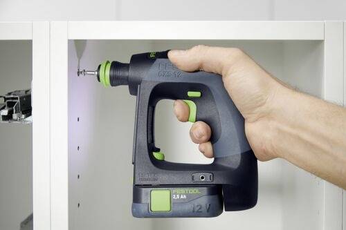 Une personne utilise un tournevis sans fil bleu foncé et vert pour visser une vis dans un meuble blanc, illustrant un assemblage de bricolage.