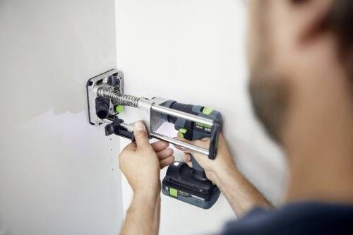 Une personne utilise une perceuse électrique pour installer une charnière métallique de porte sur une surface blanche, en se concentrant sur la précision et l'alignement.