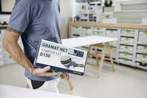 Une personne tient une boîte 'Granat Net Starter Set D150' dans un atelier. L'arrière-plan montre des étagères avec des unités de rangement et une table de travail.