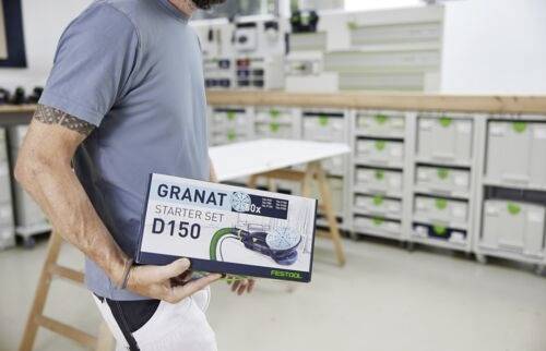 Une personne dans un atelier tenant une boîte de 'Granat Starter Set D150', avec des étagères de rangement et une table de travail en arrière-plan.