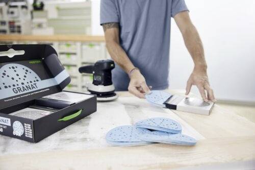 Une personne, dans un environnement d'atelier, dispose des disques de ponçage bleus étiquetés « GRANAT » à côté d'une machine de ponçage sur un établi.