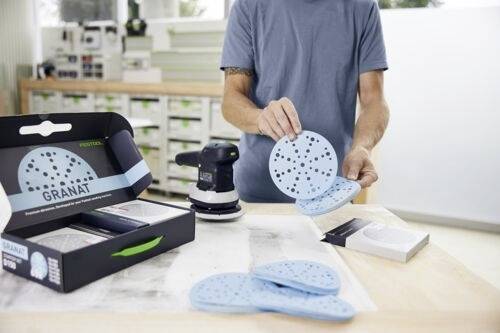 Une personne dispose plusieurs disques de ponçage bleus près d'une ponceuse sur un établi dans un atelier. Une boîte étiquetée « Granat » est posée sur la table.