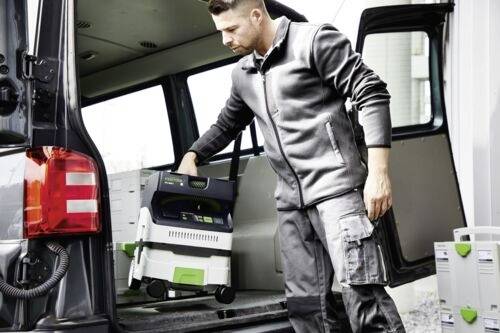 Un homme portant une veste et un pantalon gris charge un extracteur de poussière portable à l'arrière d'un véhicule, entouré de boîtes d'équipement empilées.