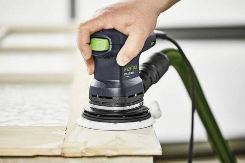 Une main tient une ponceuse électrique Festool qui lisse les bords d'un bois, avec des touches vertes visibles. L'accent est mis sur l'utilisation de l'outil et le savoir-faire artisanal.