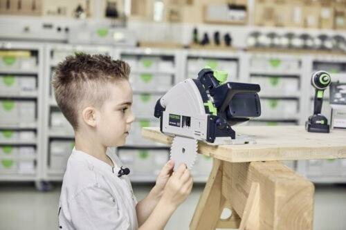 Un jeune enfant examine un outil électrique sur un établi en bois dans un environnement d'atelier, en se concentrant sur un accessoire de lame de scie circulaire.
