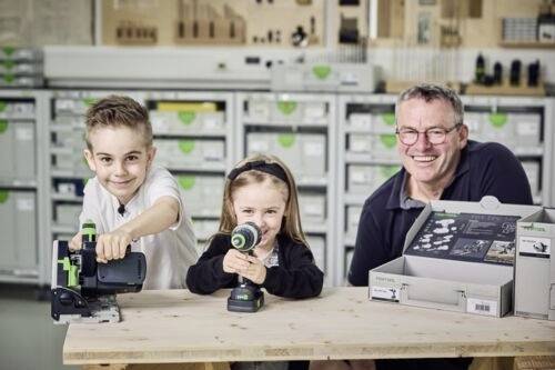 Un homme et deux enfants sourient tout en utilisant des outils électriques sur une table d'atelier. Des étagères avec des boîtes et des outils sont visibles en arrière-plan.