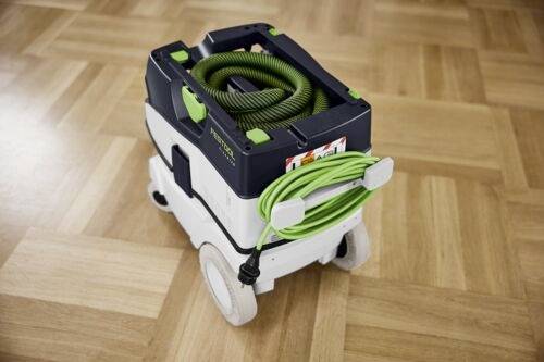 Un aspirateur compact et portable sur un sol en bois. Il dispose d'un tuyau vert enroulé sur le dessus et des touches vertes, conçu pour un transport facile.