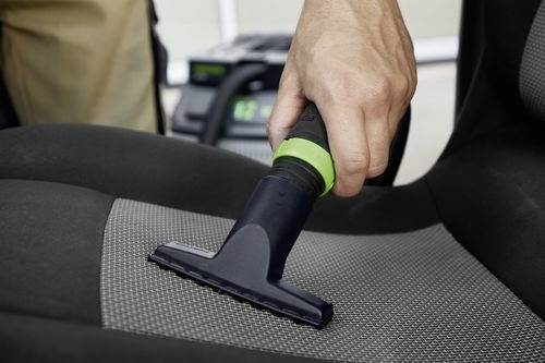 Une personne utilise un aspirateur portable avec un accent vert pour nettoyer un siège de voiture, en se concentrant sur l'élimination des saletés du tissu.