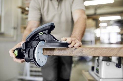 Une personne utilise une ponceuse de finition sur le bord d'une planche en bois, en se concentrant sur la précision et le lissage de la surface dans un environnement d'atelier.
