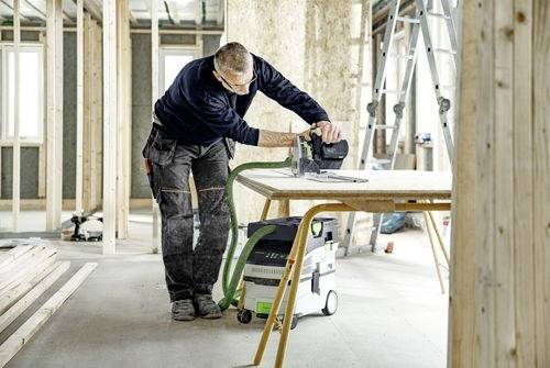 Une personne vêtue de vêtements de travail utilise un outil électrique sur une table en bois dans une pièce non finie, suggérant un projet de construction en cours.