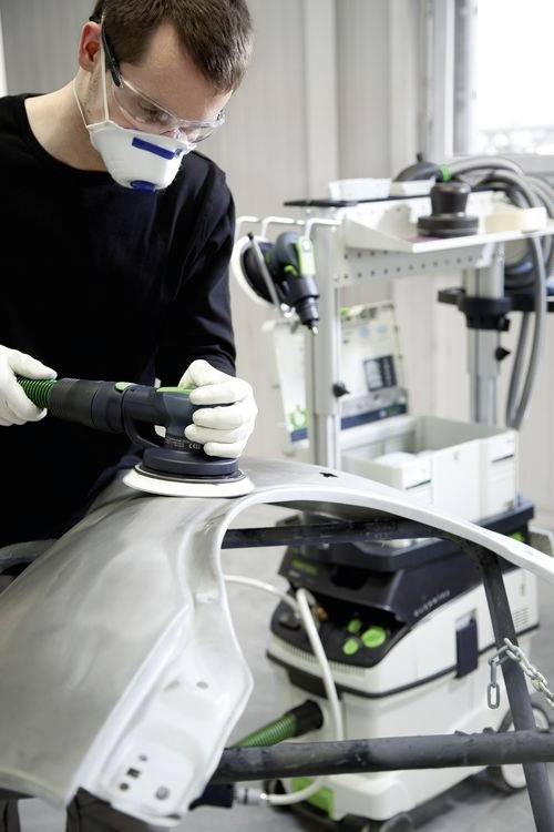 Une personne portant un équipement de protection polit une pièce automobile dans un atelier, entourée d'outils et d'équipements, concentrée sur un travail détaillé.