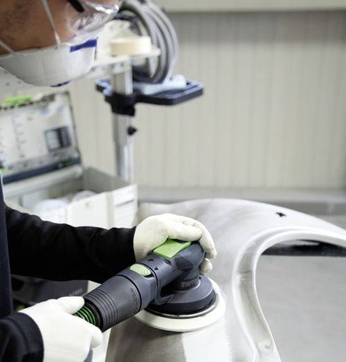 Une personne portant un équipement de protection utilise un outil électrique pour polir un pare-chocs de voiture dans un environnement d'atelier.