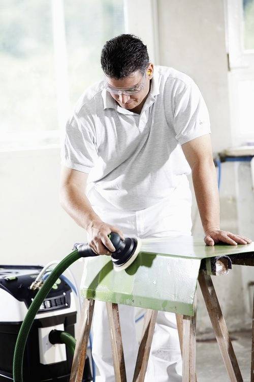 Une personne portant des lunettes de sécurité et des vêtements de travail est en train de poncer une planche en bois verte avec une ponceuse électrique dans un environnement d'atelier.