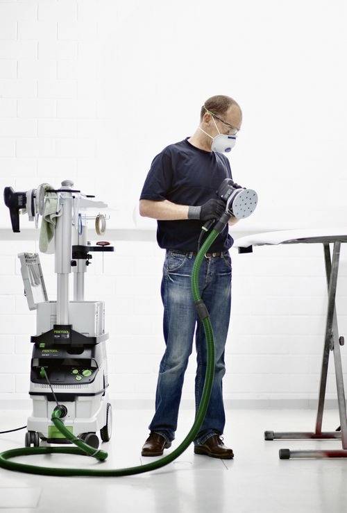 Une personne portant un équipement de protection individuelle utilise une machine à poncer, en lissant une surface. L'équipement et l'espace de travail suggèrent un environnement d'atelier.