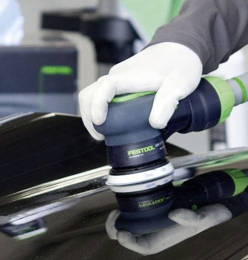 Une personne portant des gants blancs utilise un polisseur Festool pour faire briller une surface noire, reflétant le tampon de polissage et le logo.