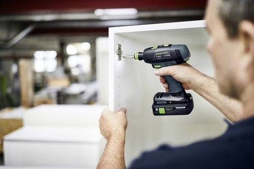 Une personne utilise une perceuse électrique pour fixer une charnière à un meuble blanc, à l'intérieur d'un atelier.