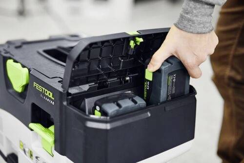 Une personne insère une batterie dans un extracteur de poussière Festool. L'appareil présente un design noir et vert, avec des compartiments pour les outils.