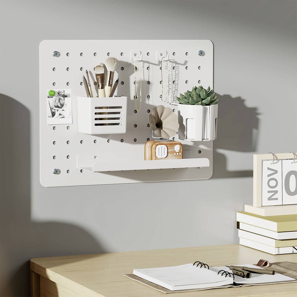 Un panneau perforé blanc accroché au mur supporte des articles de papeterie dans des gobelets, une petite plante en pot et une photo. Un agenda ouvert et un cahier sont posés sur le bureau.