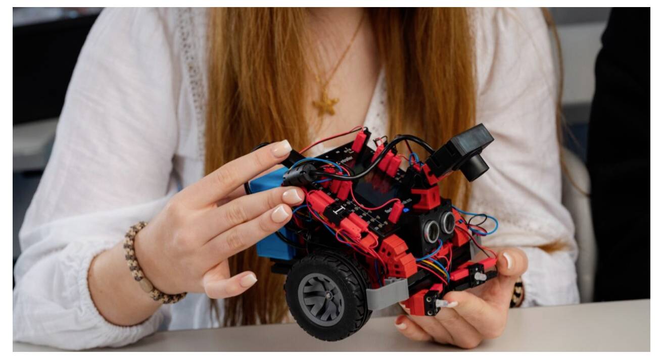 Une personne tenant un petit véhicule robotique rouge avec divers composants électroniques et des roues, présentant un projet de robotique.