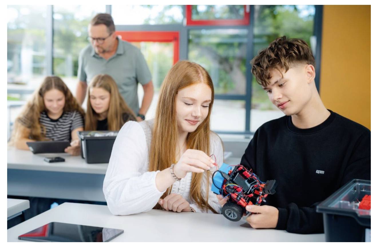 Les élèves construisent de manière collaborative un petit robot dans une salle de classe, avec un enseignant et des camarades travaillant en arrière-plan.