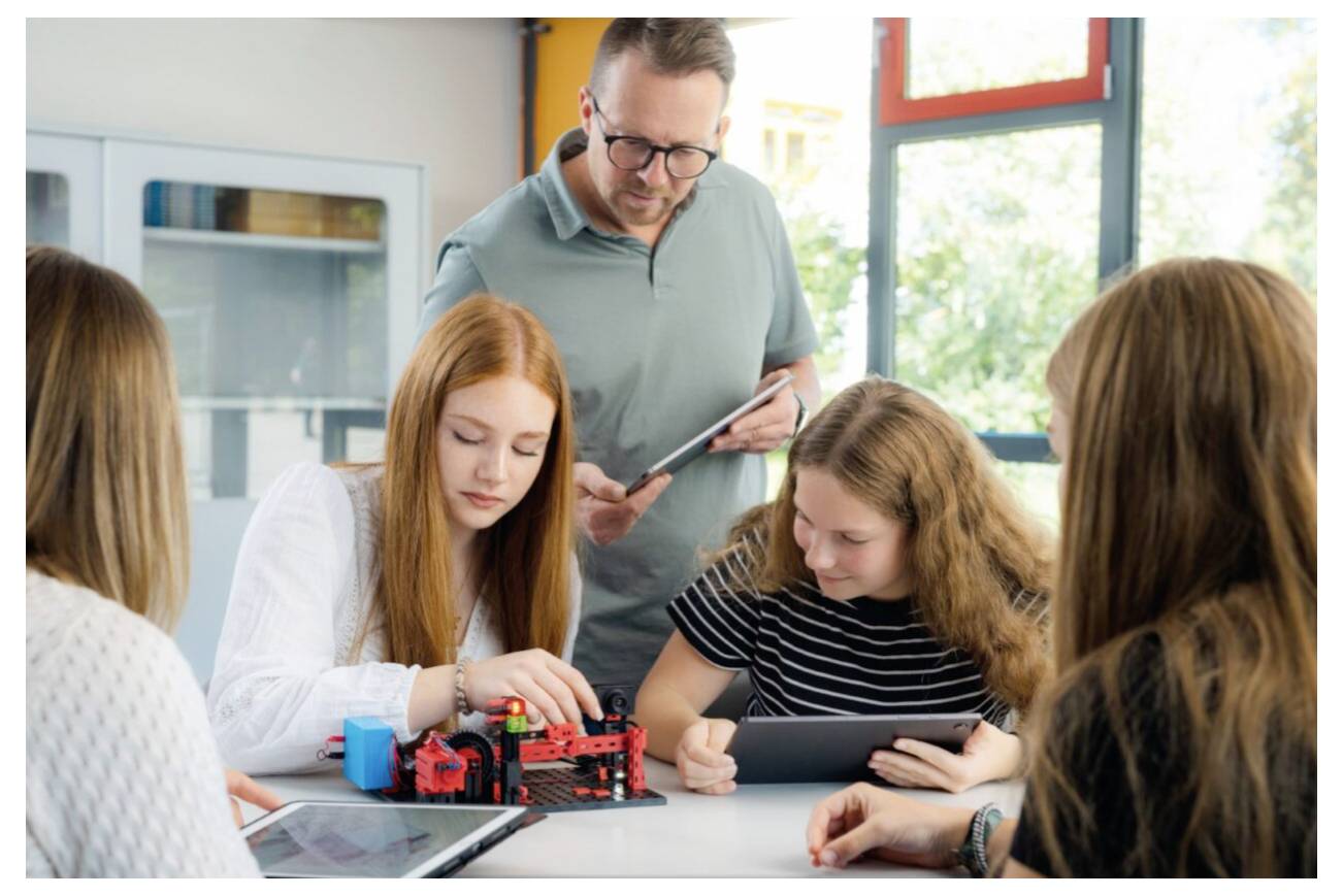 Un enseignant guide des élèves alors qu'ils travaillent sur un projet de robotique, utilisant des tablettes et un modèle de robot, dans un environnement de classe lumineux.