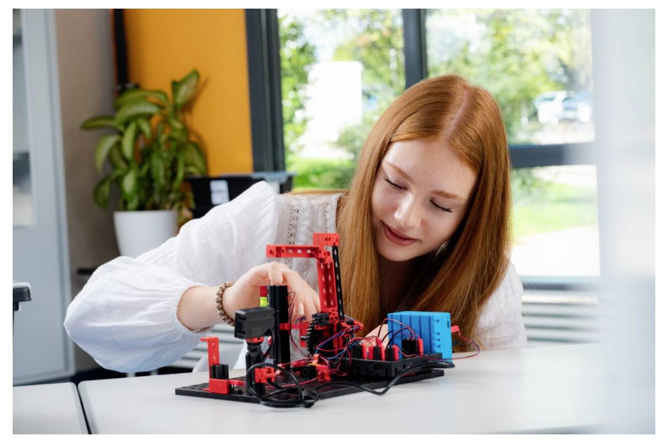 Une jeune femme aux longs cheveux roux travaille sur un petit projet de robotique, avec divers composants et fils rouges et noirs posés sur une table.
