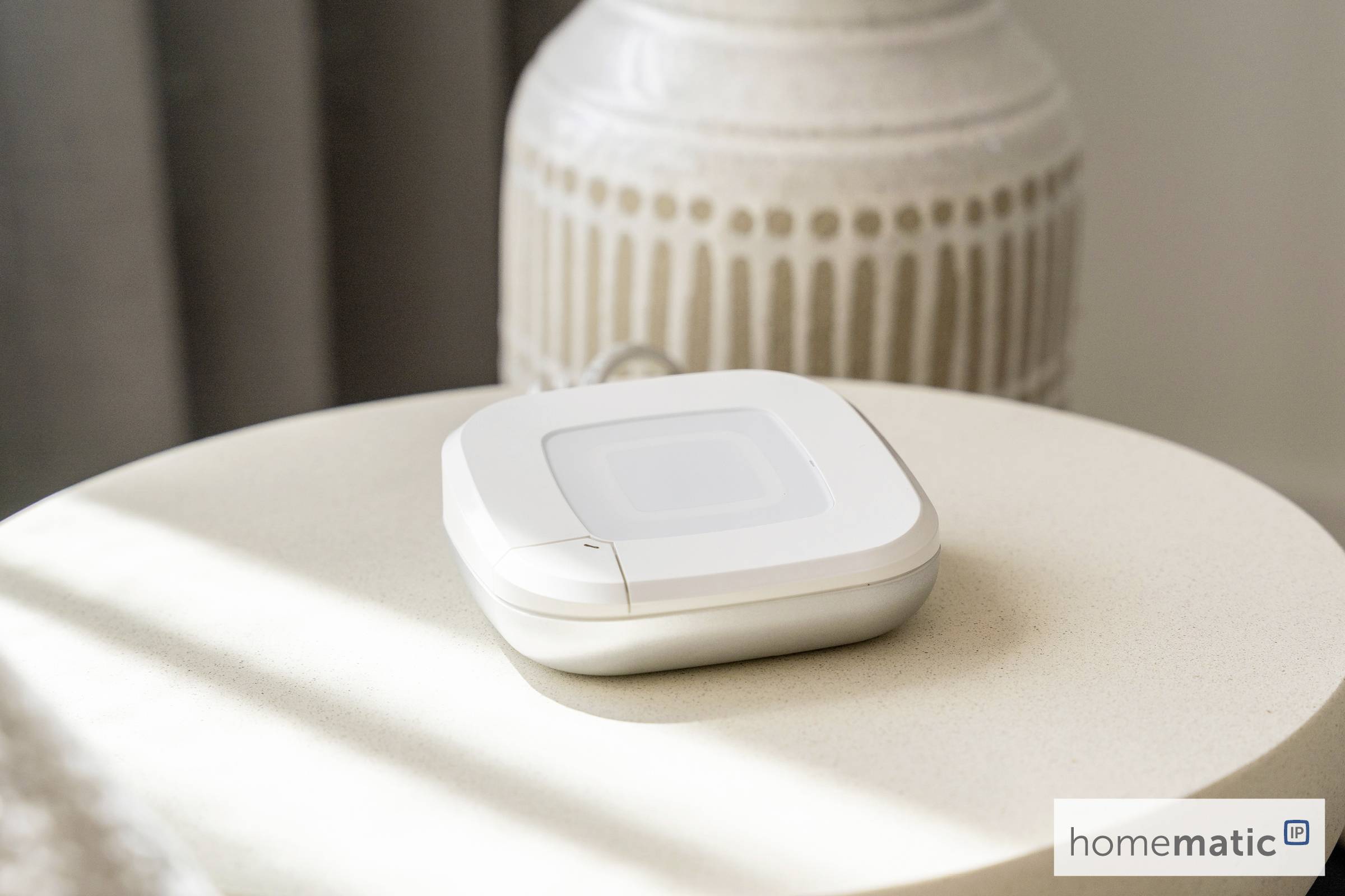 Un petit appareil domotique blanc est posé sur une table ronde beige dans une pièce doucement éclairée. Un logo indiquant 'homematic' est visible dans le coin.