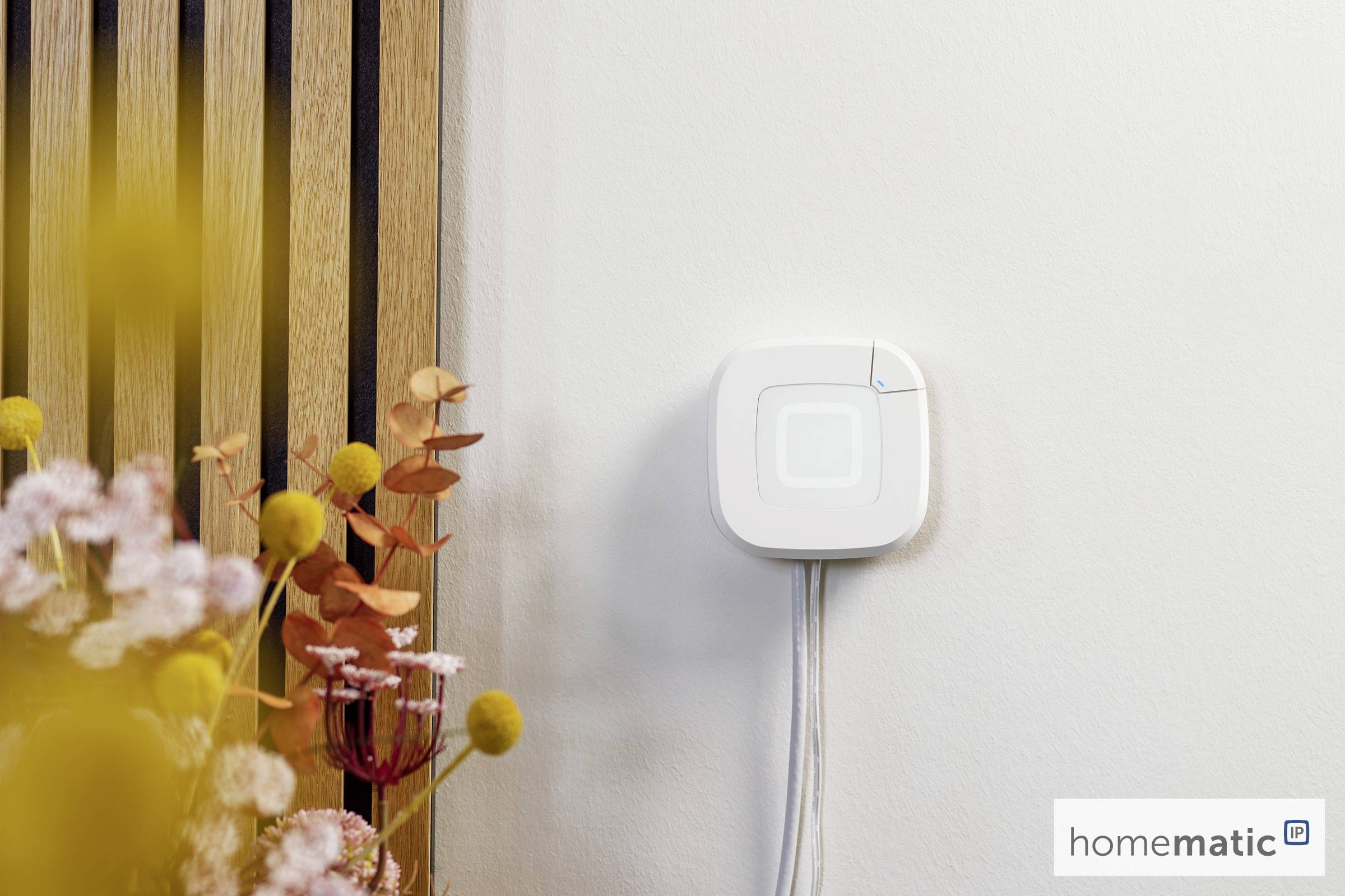 Un appareil domotique sans fil installé sur un mur à côté de fleurs décoratives, avec un voyant lumineux.