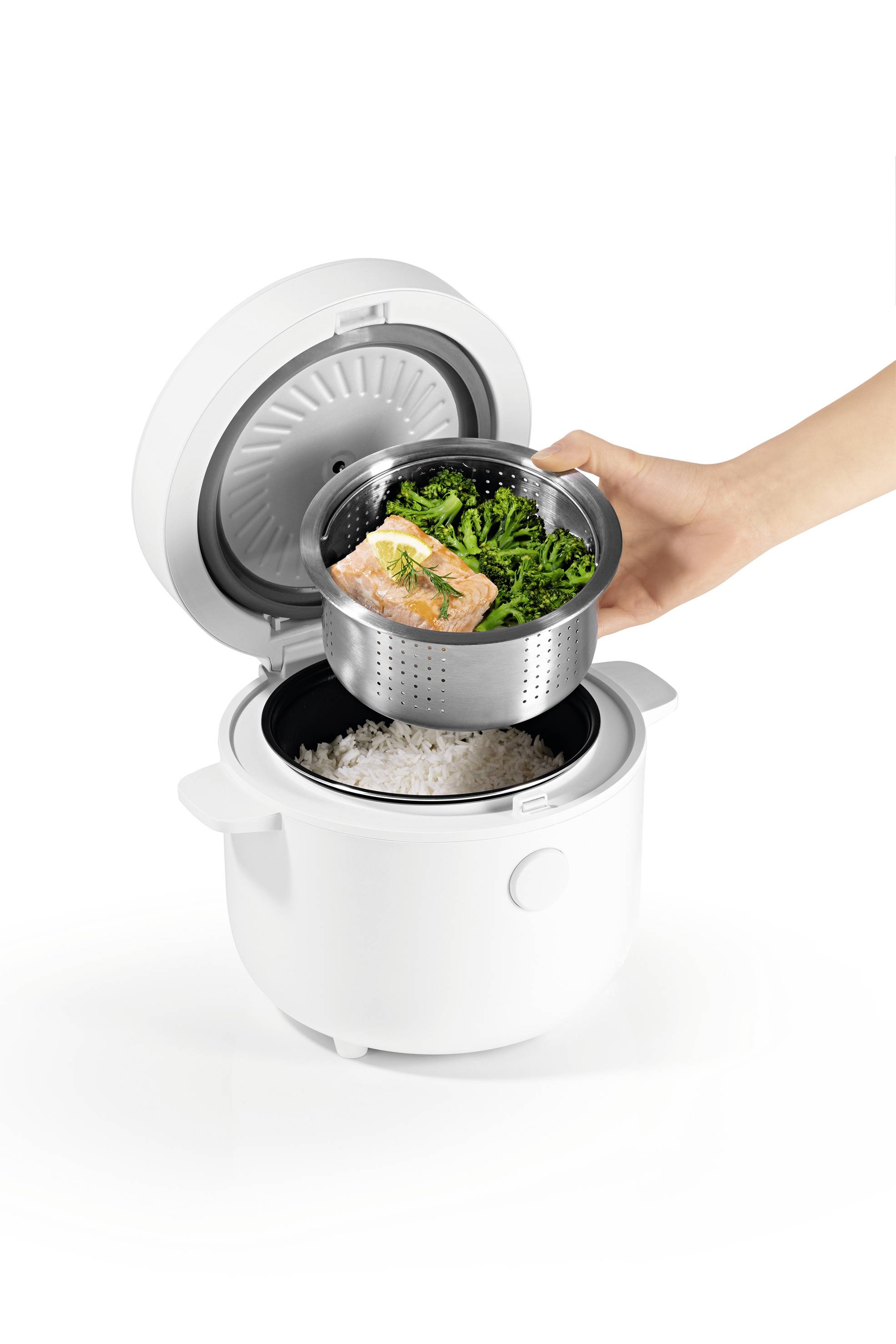 Une personne place un plateau fumant avec du saumon cuit et des légumes verts dans un cuiseur à riz, qui contient déjà du riz à l'intérieur.