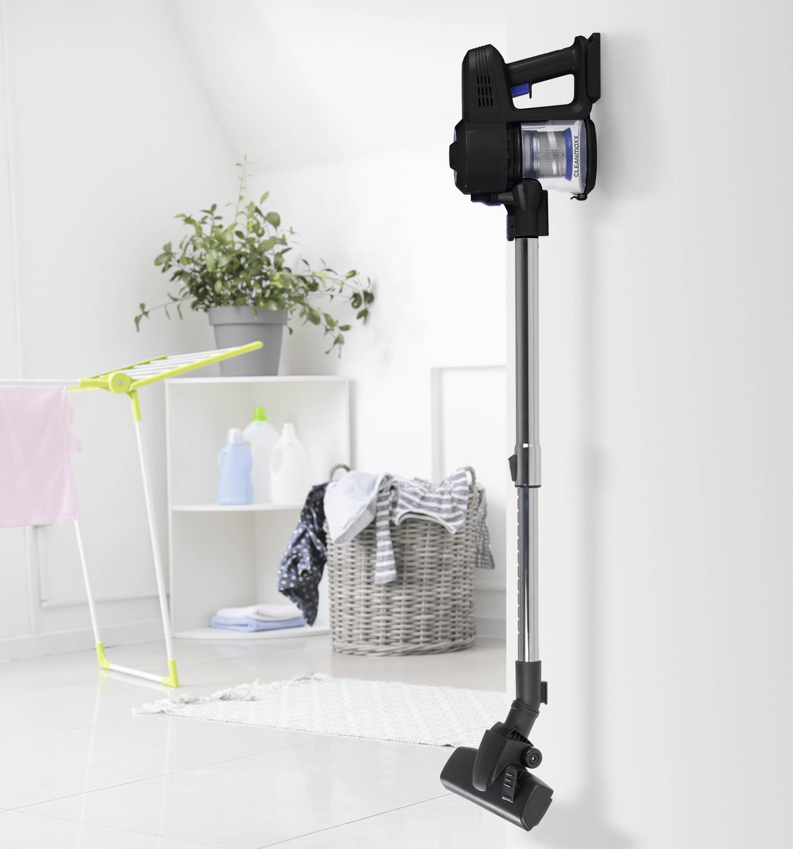 Un aspirateur sans fil fixé au mur dans un espace de vie lumineux et organisé, avec un panier à linge, un étendoir et un pot de plante à proximité.