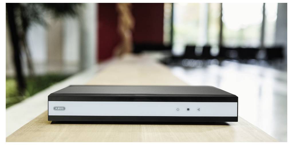 Un appareil électronique noir, probablement un lecteur multimédia ou un modem, est posé sur une surface en bois dans une pièce moderne, bien éclairée et dotée de grandes fenêtres.