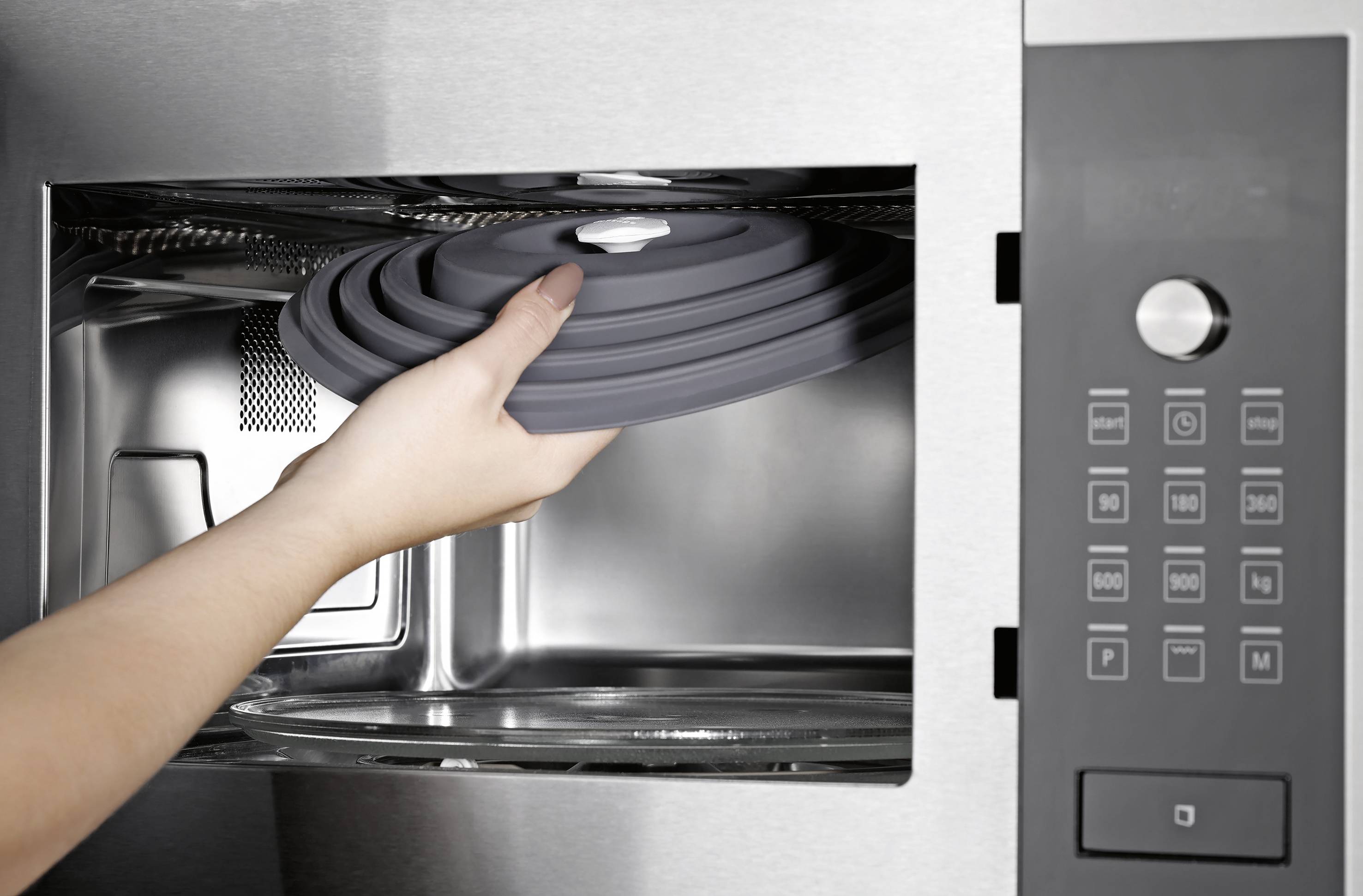 Une personne insère un couvercle en silicone dans un four à micro-ondes en acier inoxydable, en préparation pour couvrir un plat destiné à la cuisson ou au réchauffage.