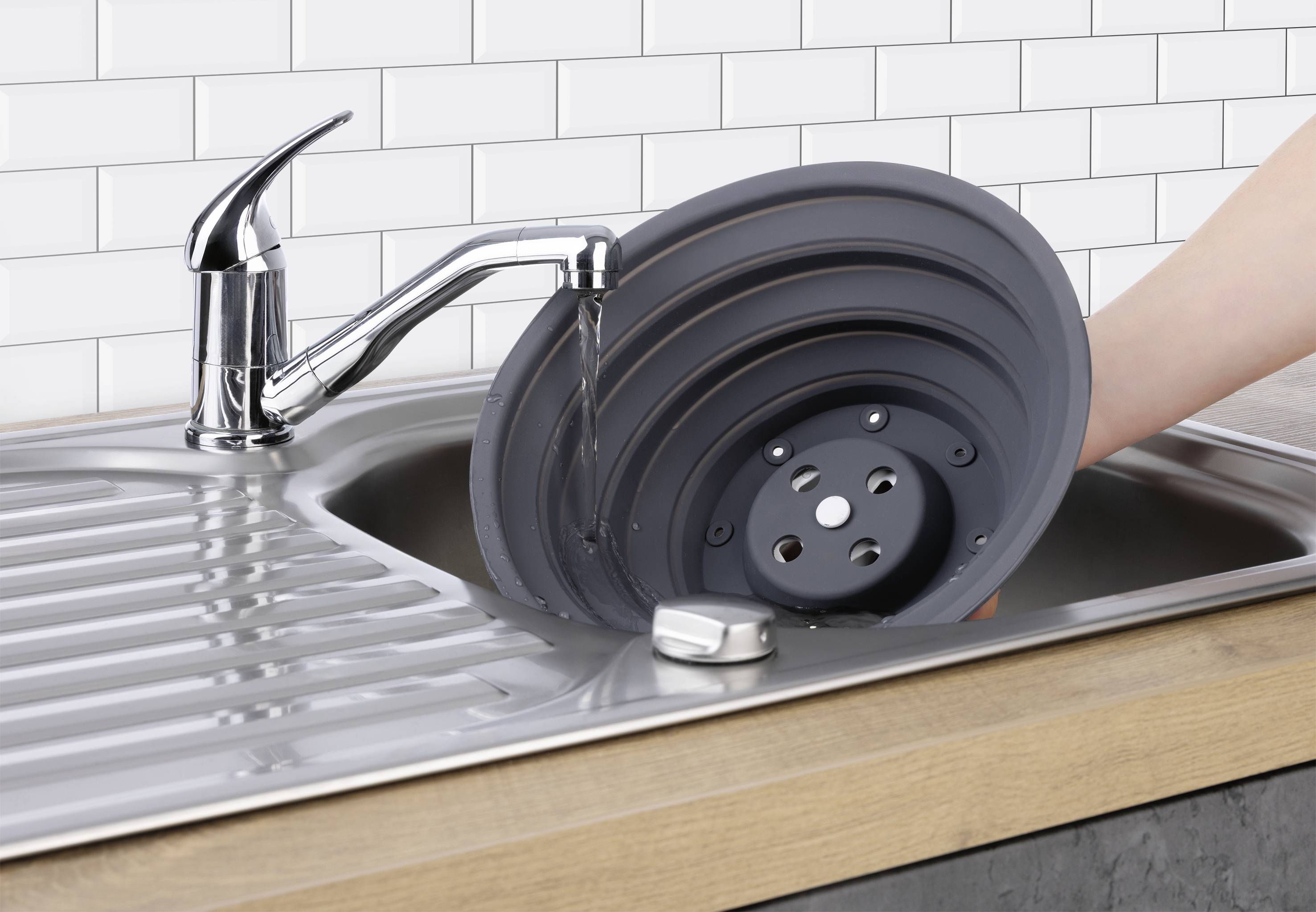 Une main lavant un égouttoir rond et pliable noir sous un robinet de cuisine avec de l'eau coulante, au-dessus d'un évier en acier inoxydable.