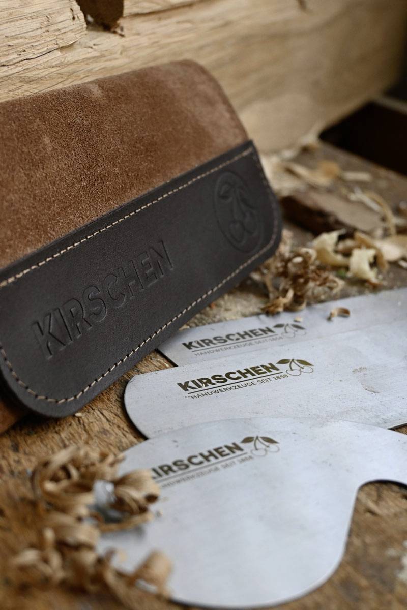 Wooden surface with wood shavings and a leather case embossed with 'Kirschen'. Two metal Kirschen tools partially visible.