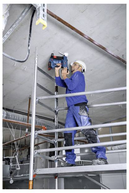 Un ouvrier portant des combinaisons bleues et un casque de chantier se tient sur un échafaudage, utilisant un outil électrique pour percer un plafond dans un environnement de construction.